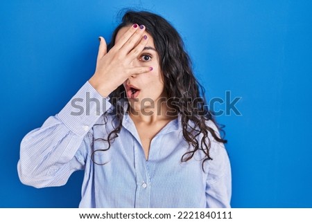 Young brunette woman standing over blue background peeking in shock covering face and eyes with hand, looking through fingers with embarrassed expression. 
