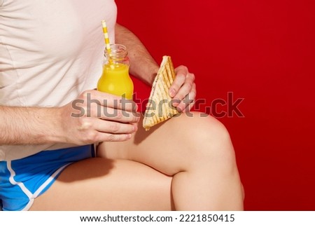 Brunch time. Female body with male hands holding sandwich and orange juice over red background. Colorful minimalism. Copy space for ad, text. Traditions and weird things