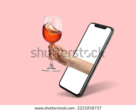 Female hand holding glass of splashing wine through screen of mobile phone on pink background Royalty-Free Stock Photo #2221858737