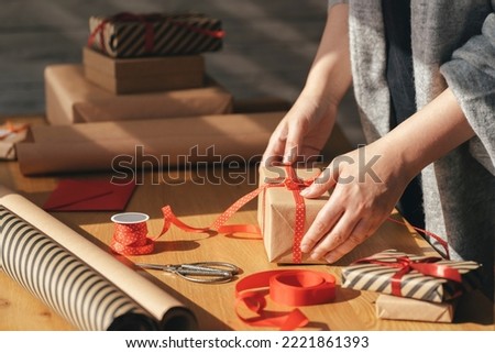 Gift wrapping in eco packaging. Young woman making bow on kraft gift box with red thread on a wooden table. Packaging and preparation of gifts for the celebration. Royalty-Free Stock Photo #2221861393