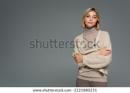 young blonde woman in turtleneck and cardigan standing with crossed arms isolated on grey Royalty-Free Stock Photo #2221880231