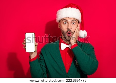 Photo of cute shocked man wear x-mas bowtie green cardigan hand arm cheek modern device empty space isolated red color background