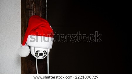 White CCTV camera in red Santa Claus hat. Outdoor video surveillance. Security system. Copy space. Web cam street control. Distance watch. Smart tech. Modern technologies. New year holiday concept. Royalty-Free Stock Photo #2221898929
