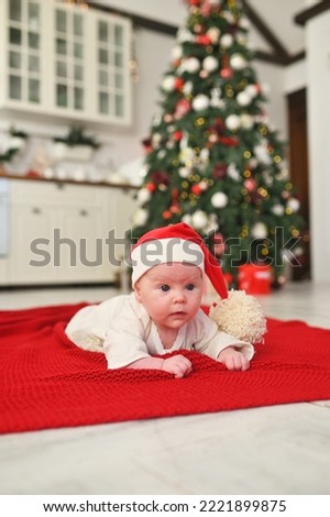 a baby in a santa hat with a large bubo lies on a red plaid against the background of a Christmas tree.
