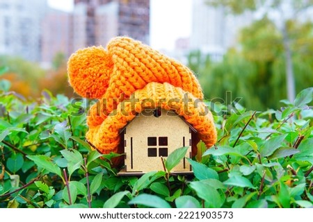 Massive winter hat put on a miniature wooden house placed on a green bush. Energy efficient heating for a country house. Royalty-Free Stock Photo #2221903573