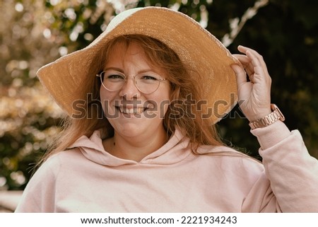 Portrait picture of a smiling woman wearing a yellow sun hat; the woman is looking straight into the camera; in the background there is a green garden