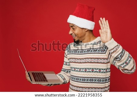 Merry young IT man wear warm cozy Christmas sweater Santa hat posing hold use work on laptop pc computer waving hand isolated on plain red background. Happy New Year 2023 celebration holiday concept