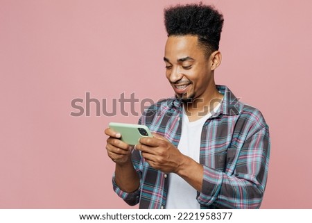 Young gambling man of African American ethnicity he wear blue shirt using play racing app on mobile cell phone hold gadget smartphone for pc video games isolated on plain pastel light pink background