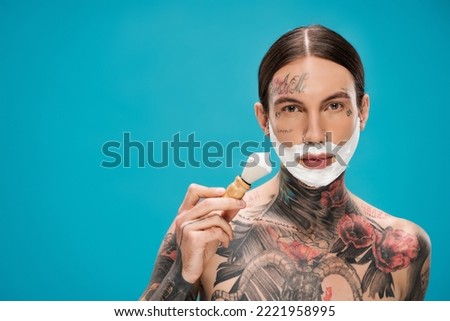 tattooed and shirtless man with shaving foam on face holding vintage brush isolated on blue.Translation of tattoo:'kiss passionate and bite gently'