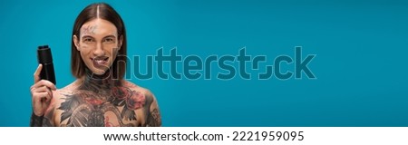 happy and tattooed young man holding antiperspirant isolated on blue, banner.Translation of tattoo:'kiss passionate and bite gently'