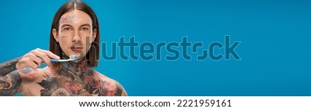 young and tattooed man holding toothbrush isolated on blue, banner.Translation of tattoo:'kiss passionate and bite gently'