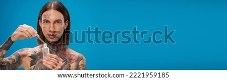 young and tattooed man holding bottle with moisturizing serum isolated on blue, banner.Translation of tattoo:'kiss passionate and bite gently'