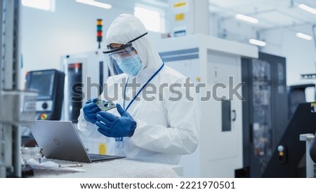 Electronic Manufacturing Factory: Engineer in Sterile Coverall Working on Laptop Computer, Examining a Circuit Board with Microchips and Testing New Electrical Equipment. Royalty-Free Stock Photo #2221970501