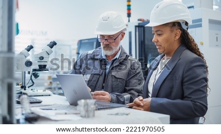 Two Professional Heavy Industry Engineers Wearing Safety Uniform and Hard Hats Discussing Industrial Machine Part on Laptop Computer. African American Specialist and Middle Aged Technician at Work.