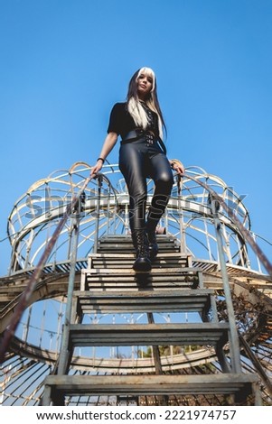 Young and skinny model with long blonde and black hair and black clothes and makeup posing over an old abandoned greenhouse with blue sky