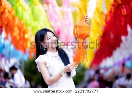 Asian woman wear white dress smiled happy and holding Lanna hanging lamp. Young woman pray bless the holy things and holding a traditional colorful paper lantern during Yi Peng lantern festival.