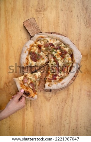 Top view of a woman's hand serving a slice of pizza made with tomato sauce, mozzarella cheese, crispy bacon and onions, in a wooden background.