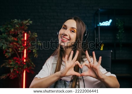 Positive young woman in headphones showing heart and smiling against the background of dark room with red backlight.