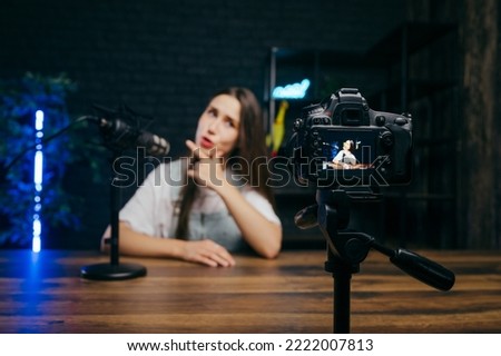 Funny woman sitting at a table and recording videos on camera with a funny face in a professional studio. Focus on the camera.