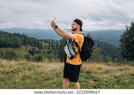 A bearded young man in casual clothes and with a backpack stands in the mountains with a smartphone in his hand and looks at the hiking route via the mobile Internet.