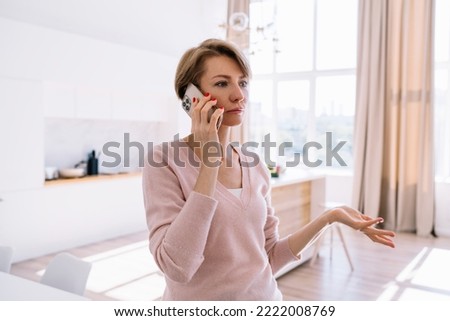 Side view of calm middle aged woman standing near table while speaking on cellphone and gesticulating with hand in sunlit room