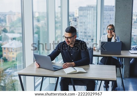 Attractive business man of Caucasian appearance in a black shirt sits at a desk using a laptop in a modern office with colleagues in the background. Working atmosphere in the office.