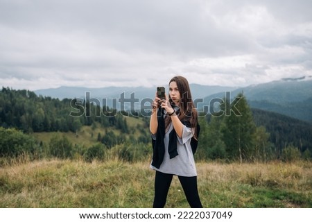 A beautiful woman with a smartphone in her hands stands in the mountains against the background of beautiful Carpathian views and takes a photo on the smartphone camera.