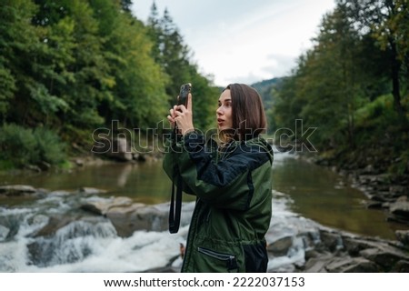 Surprised female tourist in a jacket stands on the river bank in the mountains on a rock and takes a photo on the smartphone camera against the background of the forest and beautiful views.