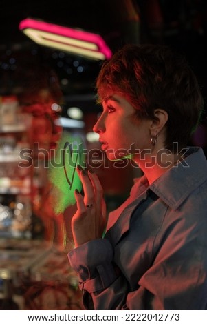 Side view of sad lonely millennial girl with short haircut standing in red neon light on street at night and drawing heart with finger on shop window, missing loved one. Dreaming about falling in love