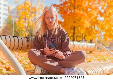 a blonde schoolgirl sits on a bench and looks at the phone against the background of yellow autumn trees.