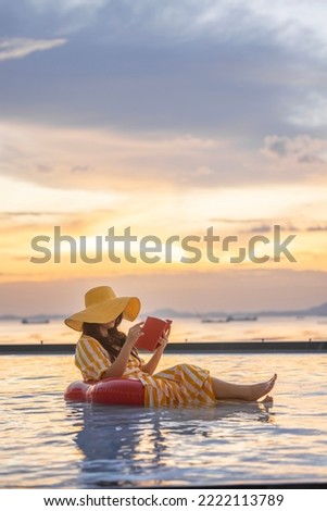Vertical photo of Happy tourist woman in casual clothes hat, reading a book sitting on red inflatable ring, relaxing by the pool in a luxurious beachfront hotel at sunset enjoying beach vacation. Royalty-Free Stock Photo #2222113789