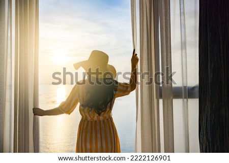 Rear back view woman opening curtains, relaxing and looking beautiful seascape panorama with rays of sun light in holiday on the balcony in high quality hotel feeling happy. Composition of nature Royalty-Free Stock Photo #2222126901
