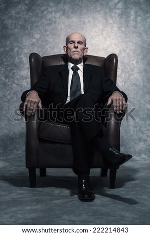 In leather chair sitting senior businessman with gray beard wearing dark suit and tie. Against grey wall. Royalty-Free Stock Photo #222214843