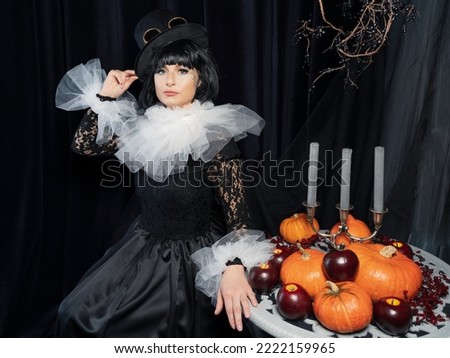 A beautiful brunette in a vintage black dress, a white frill and a vintage hat is sitting at a table with pumpkins and candles. The concept of a happy Halloween holiday.