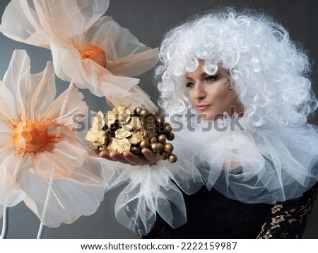 A beautiful girl in a vintage black dress, a white frill and a lush curly wig poses in a studio with huge flowers. Carnival costume.