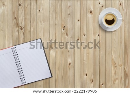 Top view picture of clear white coffee cup with smiling face from coffee bubbles and empty notebook put on wooden table floor. Background for flat lay design or minimalist style and copy space.