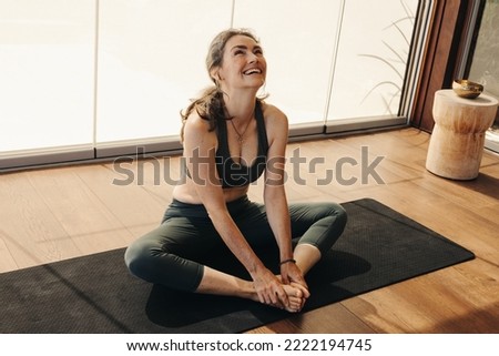 Senior woman smiling cheerfully while doing a butterfly pose on a yoga mat. Mature woman practicing a relaxation exercise at home. Happy senior woman having a session of hatha yoga. Royalty-Free Stock Photo #2222194745