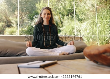 Happy holistic healer smiling at the camera while meditating in her living room. Spiritual senior woman practicing hatha yoga at home. Healthy mature woman practicing a mindful and zen lifestyle.