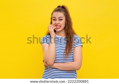 Insecure young woman biting fingernails, looking scared, trembling fear, watching something scary, standing anxious in white-blue striped t shirt over yellow background Royalty-Free Stock Photo #2222200881