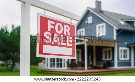 Close Up of a Red Real Estate Property for Sale Flopping in the Wind. Background with a Big Blue Stylish Countryside House. Housing Market Concept with Residential Home on Sale in the Suburbs.
