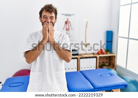 Young handsome physiotherapist man working at pain recovery clinic laughing and embarrassed giggle covering mouth with hands, gossip and scandal concept  Royalty-Free Stock Photo #2222219329