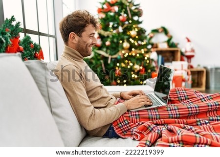 Young hispanic man using laptop sitting by christmas tree at home