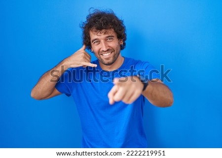 Hispanic young man standing over blue background smiling doing talking on the telephone gesture and pointing to you. call me. 