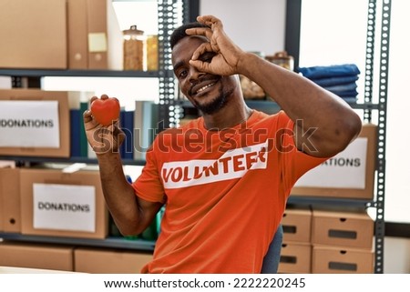 Young african man wearing volunteer t shirt at donations stand holding heart smiling happy doing ok sign with hand on eye looking through fingers 