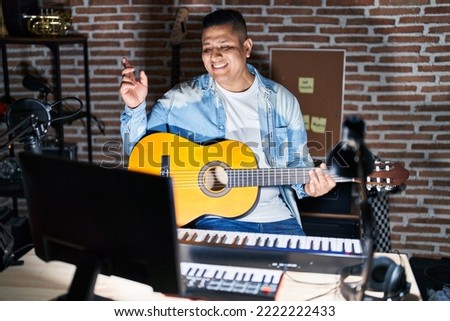 Hispanic young man playing classic guitar at music studio with a big smile on face, pointing with hand finger to the side looking at the camera. 