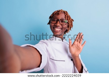 Close up shot of african american chef holding camera to shoot a selfie. Optimistic female professional chef looking at camera with big smile against blue background, studio shot.