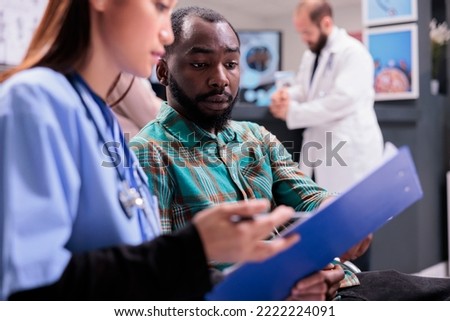 Close up of african american man reviewing medical records along with asian female member of in house hospital staff. Doctor indicating treatment to young male patient at sanatorium lobby. Royalty-Free Stock Photo #2222224091