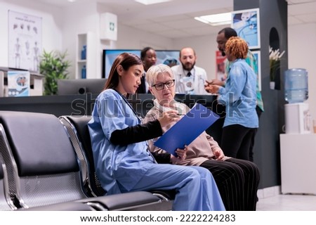Asian gerontology physician in charge of gerontology area requesting personal data from elderly patient. Medical clinic receptionist personnel attending african american sick young couple of people. Royalty-Free Stock Photo #2222224283