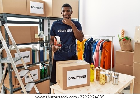 Young african american volunteer man packing donations box for charity with hand on chin thinking about question, pensive expression. smiling and thoughtful face. doubt concept. 