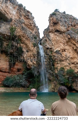 Beautiful vertical landscape with two people and the back of a dog in the foreground of a waterfall in the cave de turches surrounded by vegetation and rocks in Buñol, Valencia, Spain